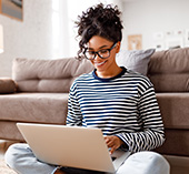 Woman with glasses sitting next to couch smiling at laptop