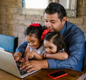 man sitting at computer with toddler daughters on lap