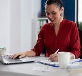 Women sitting at computer, writing in notepad with a coffee mug.