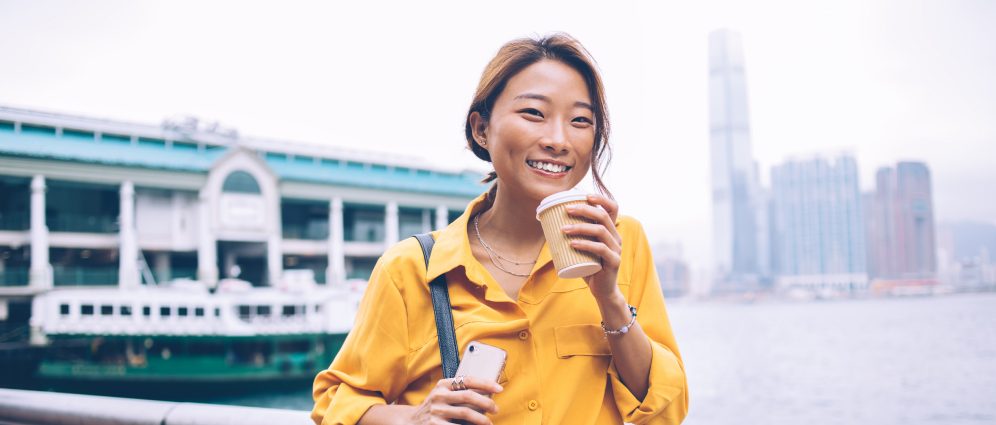 Woman walking by river in city, holding coffee and phone.